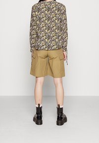 Florale Bluse mit langen Ärmeln in Schwarz, Gelb und Lila; kombiniert mit khakifarbenen Cargo-Shorts; schwarze klobige Stiefel; weiße Socken.