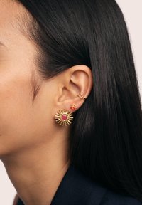 Pendientes de oro con un diseño de estallido de sol y una piedra rosa central, combinados con un pequeño pendiente rojo y un aro de oro sencillo, llevados en la oreja.