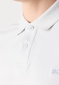 Gros plan d'un col de polo blanc avec deux boutons fermés et un texte brodé partiel bleu clair sur la poitrine.