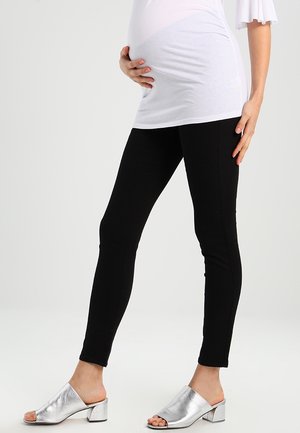 Persona embarazada con leggings negros, una camiseta blanca y zuecos plateados de tacón ancho y abierto en los dedos, posando contra un fondo liso.