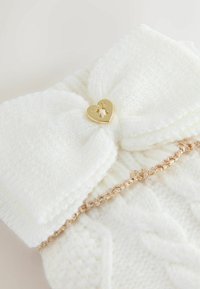 Bijeli pleteni dodatak s velikom mašnom, zlatnim srcem u obliku srca s natpisom "Baker" i zlatnim pletenim rubom duž rubova.