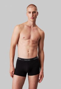Bóxer negro con una cintura de marca, hecho de un material suave y elástico. El modelo está sin camiseta, posando frente a un fondo gris.