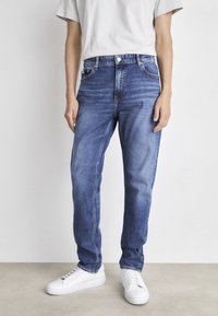 Blue denim jeans with a straight fit, featuring a button closure, five pockets, and light washing. Paired with white sneakers.