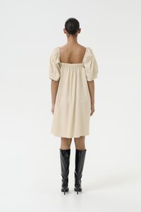 Vestido beige de mangas abullonadas con espalda cuadrada y cintura con fruncidos, combinado con botas altas de charol negro. Tejido suave y ligero.