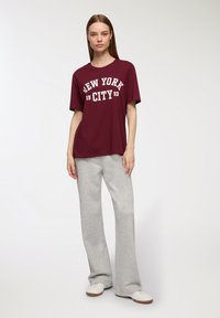 T-shirt di cotone bordeaux con la dicitura "NEW YORK CITY 93" stampata in bianco. Indossato con pantaloni sportivi grigi chiari a campana e sneakers bianche.