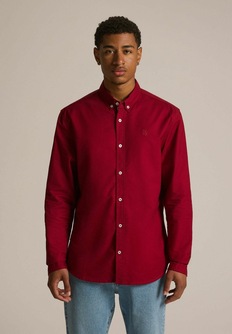 Czerwona bawełniana koszula z długim rękawem, zapinana na guziki, z zaokrąglonym dołem, białymi guzikami i subtelnym logo na piersi. Połączona z niebieskimi dżinsami.
