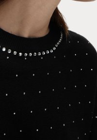 Pull noir avec un col rond, orné de strass argentés autour du col et de strass éparpillés sur l'ensemble du tissu.