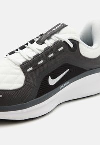 Zapatilla deportiva de Nike con parte superior sintética en blanco y negro, tejido texturizado, paneles laterales acolchados y suela de goma gris con el logo Air.
