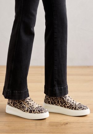 Person iført sorte jeans og leopardmønstrede lave sneakers med hvide såler, der står på et trægulv.