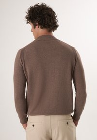 Bruine lange mouwen trui met een geribde kraag en schouderdetails, gedragen over beige broek. Glad stoftextuur, aansluitend ontwerp.
