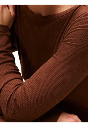 Personne portant une chemise marron moulante à manches longues côtelée, les bras croisés près de la poitrine et le cou visible.