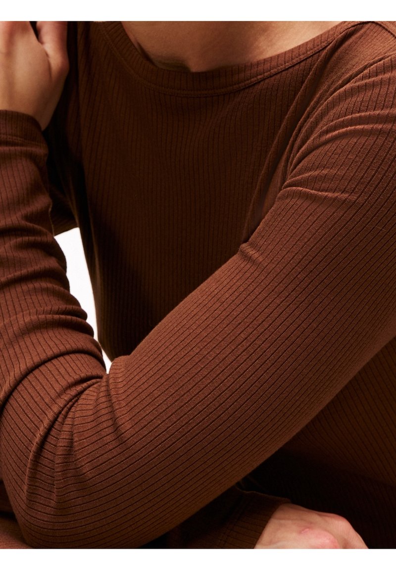 Personne portant une chemise marron moulante à manches longues côtelée, les bras croisés près de la poitrine et le cou visible.