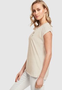 Beige bomull t-skjorte med korte, rullet ermer, rund halslinning, og blomstermønsterbroderi foran. Modell iført hvite jeans.