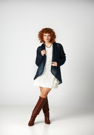 Kręconowłosa rudowłosa kobieta w białej, warstwowej sukience, granatowym otwartym kardiganie, złotym naszyjniku łańcuszkowym i brązowych kozakach za kolano stoi na białym tle.