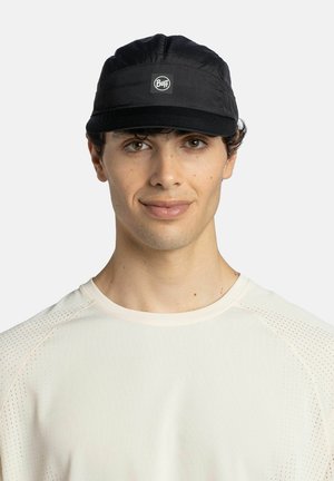 Junger Mann mit schwarzer Buff-Kappe und hellbeigem, perforiertem Kurzarmhemd, der nach vorne schaut und leicht lächelt.