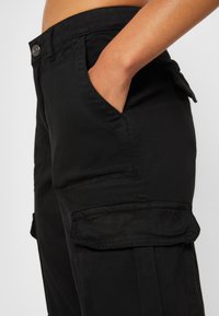 Pantaloni cargo neri realizzati in misto cotone, con tasche laterali, una texture liscia e una vita standard con chiusura a bottone.