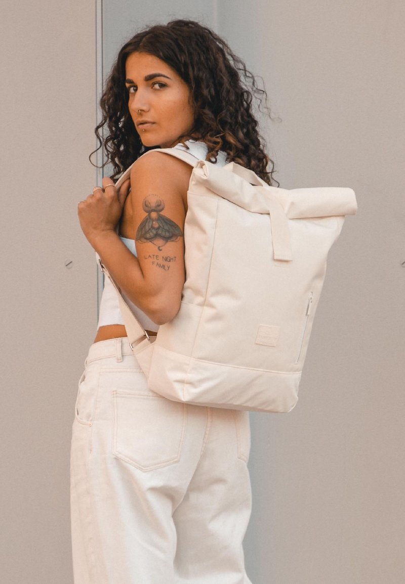 Görbe hajú nő fehér topot és nadrágot visel, egy bézs tekercses hátizsákot cipel, és a karján egy rovar tetoválás látható, mellette a "LATE NIGHT FAMILY" felirattal.