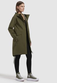 Olíva zöld esőkabát kapucnival, cipzárral elől és oldalzsebekkel; fekete leggings-szel és olíva zöld magasszárú tornacipővel párosítva.