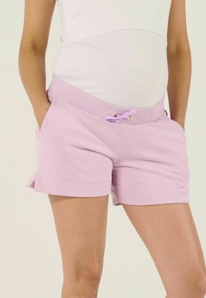Schwangere Frau trägt lila Umstandsshorts mit elastischem Bund und Kordelzug, Hände in den Taschen, sowie ein weißes eng anliegendes Oberteil.