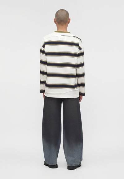 Άτομο που στέκεται γυρισμένο με την πλάτη, φοράει άσπρο πουκάμισο με μακριά μανίκια με ναυτικές και ελιάς ρίγες και φαρδιά ξεθωριασμένα μαύρα τζιν.