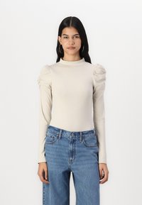 Cămașă bej cu mâneci lungi, guler înalt și m�âneci bufante, asortată cu blugi din denim albaștri, cu talie înaltă. Textură netedă, detalii minime.