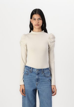 Langärmliges, beiges Oberteil mit hohem Kragen und puffigen Ärmeln, kombiniert mit blauen, hochtaillierten Jeans. Glatte Textur, minimale Details.