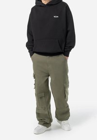 Person, der einen schwarzen Kapuzenpullover mit kleinem weißem Logo, olivegrüne Cargo-Hosen mit mehreren Taschen und weiße Sneaker trägt, steht mit den Händen in den Taschen.
