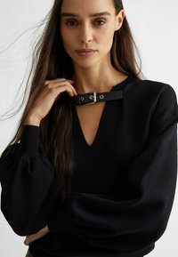 Maglione nero a maglia con scollo a V profondo e un'accento di cinturino in pelle sul colletto. Presenta maniche lunghe a sbuffo e una finitura testurizzata.