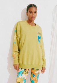 Lichtgroene sweatshirt met lange mouwen, featuring een blauwe hartgrafiek en kleurrijke ontwerpen langs de mouwen; relaxte pasvorm. Kleurrijke gestreepte leggings.