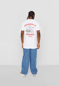 Weißes Baumwoll-T-Shirt mit einer roten Grafik eines Kochs, der eine Pizza hält, mit "Giuseppe's Pizzeria" und der Kontaktnummer darunter aufgedruckt.