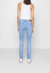 Vaqueros de mezclilla azul claro con un corte ajustado, que cuentan con bolsillos traseros y un parche de logo marrón. Combinados con zapatillas blancas y una blusa sin mangas.