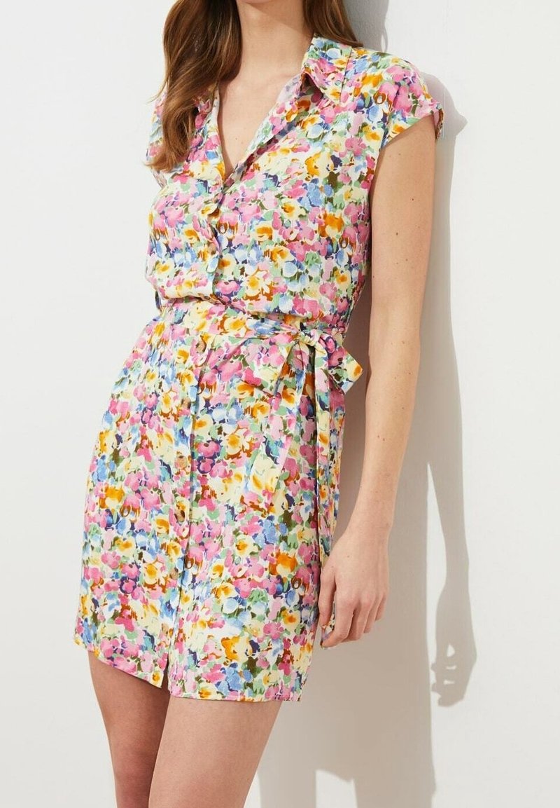 Femme portant une robe fleurie à manches courtes avec ceinture, aux fleurs roses, jaunes, bleues et blanches, debout contre un mur blanc.