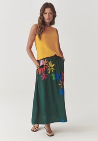 Top galben texturat fără mâneci, asortat cu o fustă lungă verde cu modele florale colorate și buzunare laterale. Flip-flops negre.