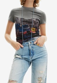 Grå kortærmet T-shirt med farverigt grafisk print; afslappet pasform. Lys blå slidte jeans med huller og dekorative syninger.