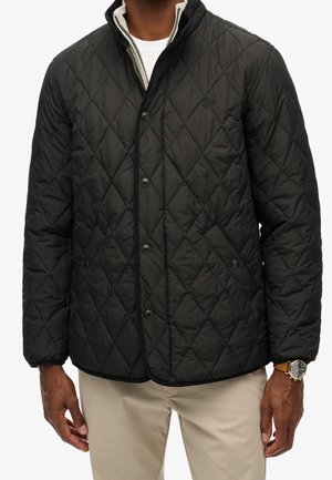 Hombre que lleva una chaqueta negra acolchada con botones a presión, pantalones beige, camisa blanca y un reloj con correa de cuero marrón y esfera negra redonda.