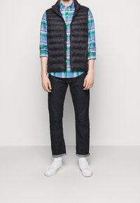 Muž ve černé prošívané vestě přes modrou a zelenou kostkovanou košili, tmavých zkrácených džínech, bílých ponožkách a bílých teniskách na jednobarevném pozadí.