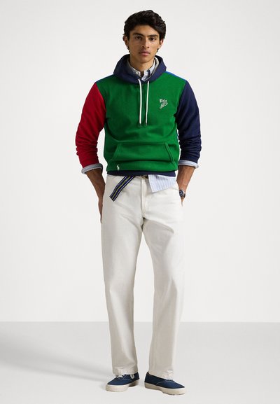 Color-blocked hoodie in green, red, and navy with a kangaroo pocket. Paired with cream trousers and navy sneakers. White shirt underneath.