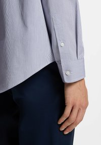 Zbliżenie na dłoń osoby spoczywającą wzdłuż ciała, ubraną w jasnoniebieską koszulę w paski z długim rękawem oraz ciemnoniebieskie spodnie.