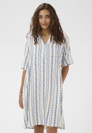 Jonge vrouw met krullend haar, gekleed in een witte jurk tot op de knie met verticale blauwe bloemstrepen en korte mouwen.