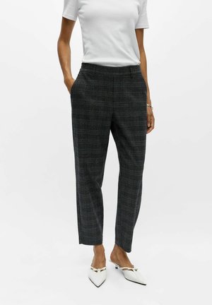 Femme portant un pantalon à carreaux gris foncé, une chemise blanche à manches courtes et des chaussures blanches pointues, debout les mains dans les poches.