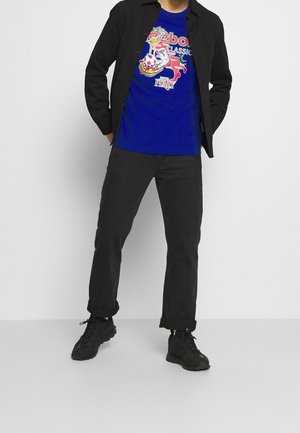 Persona con chaqueta negra, camiseta gráfica azul con texto de Italia y Reebok, pantalones negros y zapatillas negras, de pie con las manos en los bolsillos.