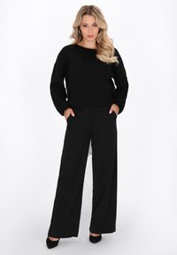 Schwarzer Strickpullover mit Rundhalsausschnitt, kombiniert mit schwarzen weiten Hosen. Die Hose hat seitliche Taschen. Auffällige schwarze High Heels runden den Look ab.