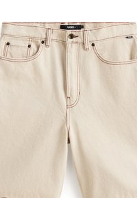 Shorts en denim beige avec des surpiqûres rouges, design à cinq poches et fermeture à bouton. Présente une étiquette logo Vans sur la ceinture.