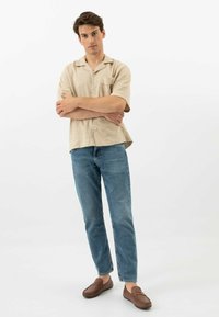 Camicia beige a maniche corte con bottoni, jeans blu e mocassini marroni. La camicia presenta una tasca singola e una vestibilità rilassata.