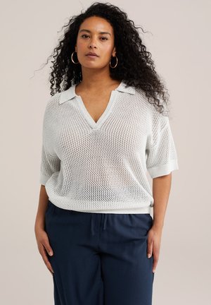 Žena s kučeravými vlasmi, ktorá nosí zlaté kruhové náušnice, biely voľný sveter s krátkym rukávom a námornícke modré nohavice so šnúrkou, stojaca pred neutrálnym pozadím.