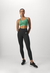 Soutien-gorge de sport vert avec des logos blancs, associé à un legging noir taille haute avec une poche. Des chaussures de sport noires complètent la tenue.