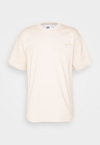 Beige t-paita lyhyillä hihoilla ja pyöreällä kaula-aukolla, jossa on pieni sävyton logo vasemmassa rinnassa, esitetty valkoista taustaa vasten.