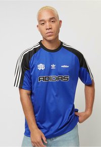 Blå Adidas-tröja med svarta axlar, vita ränder och en stor svart "ADIDAS"-logotyp. Slät tyg, sportig design, kortärmad.