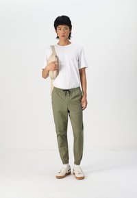 Witte t-shirt, olijfgroene joggerbroeken met elastische boorden, en beige schoenen met lichtbruine zolen. Persoon houdt een beige schoudertas vast.