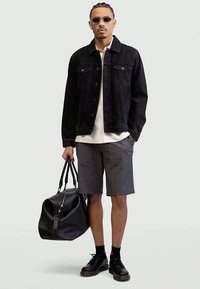 Jaqueta de gangue preta por cima de uma camisa branca, calções cinzentos às riscas, sapatos pretos e óculos de sol, segurando um saco de desporto preto.